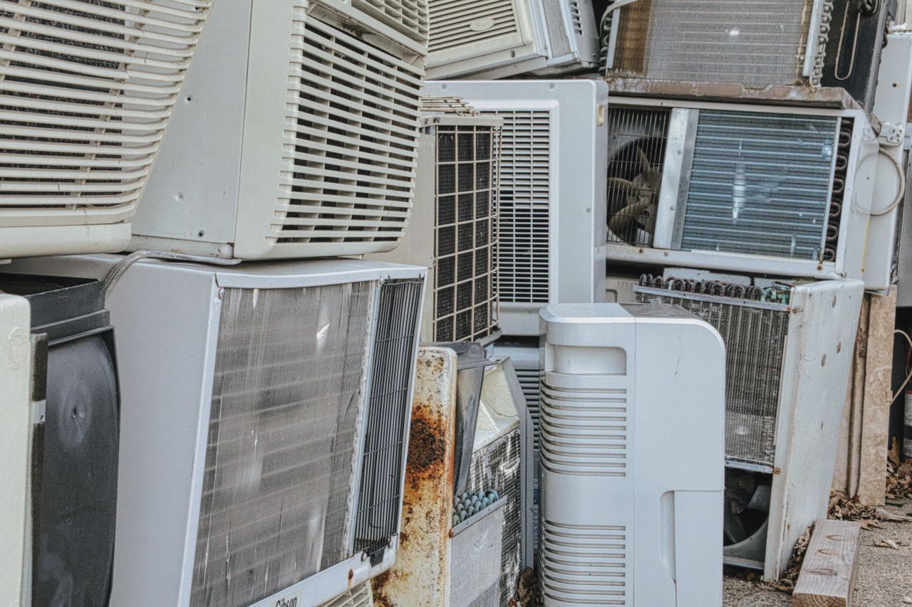 About A collection of various old and broken air conditioning units piled outdoors.